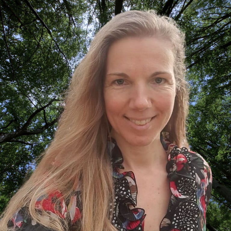 Woman with long reddish-blonde hair smiling outdoors under green tree canopy, wearing a floral patterned top